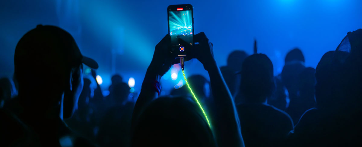 A person raises a cell phone connected to a glowing cable in front of a large crowd, capturing the moment during an event